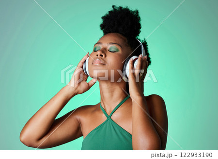 Black woman in green studio with headphones on, listening to music. Young happy girl with makeup, cosmetics and beauty with wireless earphones streaming a song, podcast or radio for fun and to relax 122933190