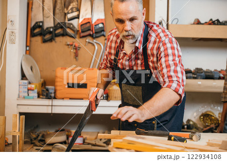 Senior carpenter in a red plaid shirt saws wood in a workshop. Experienced craftsman working with precision, focused on creating quality furniture with tools and materials in a carpentry setting Senior carpenter in a red plaid shirt saws wood in a workshop. Experienced craftsman working with precision, focused on creating quality furniture with tools and materials in a carpentry setting 122934108