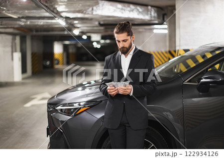 Young Caucasian male businessman counts cash in underground parking lot next to car, conveying secrecy and business. 122934126