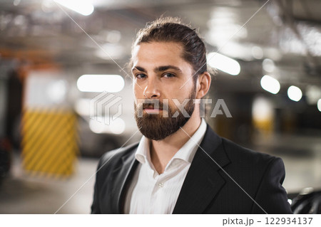 Portrait of young Caucasian male businessman in underground parking, exuding confidence and professionalism, wearing a suit with serious expression against a blurred industrial background. Portrait of young Caucasian male businessman in underground parking, exuding confidence and professionalism, wearing a suit with serious expression against a blurred industrial background. 122934137