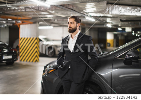 Young Caucasian man in formal suit leaning against car in underground parking lot. Business professional exuding confidence, poised in urban setting. Modern lifestyle and transportation theme. 122934142