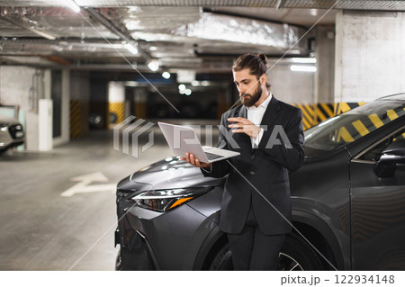 Young Caucasian male businessman in suit conducts video call on laptop in underground parking lot beside car. Business communication and remote work. 122934148