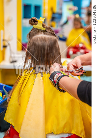 Back view of hairdresser making a hair style to cute little girl in real child salon with colorful interior and toys Back view of hairdresser making a hair style to cute little girl in real child salon with colorful interior and toys 122934286