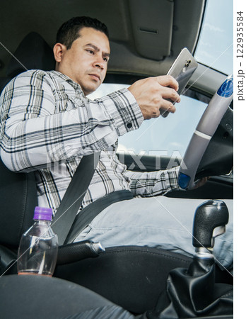 Man sitting in his car texting with his cell phone, Side view of a young man sitting inside car using mobile phone, Young man sitting in a car and looking at a mobile phone Man sitting in his car texting with his cell phone, Side view of a young man sitting inside car using mobile phone, Young man sitting in a car and looking at a mobile phone 122935584