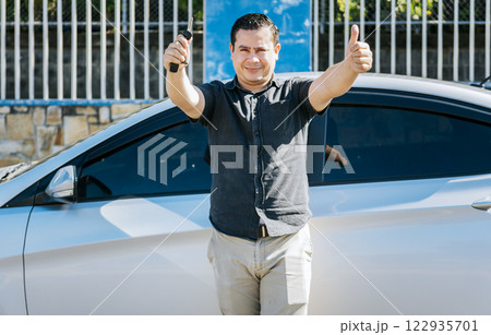 Male driver standing next to the car showing the keys with thumb up. Smiling owner with new car standing showing keys, Driver leaning on the car showing the key 122935701