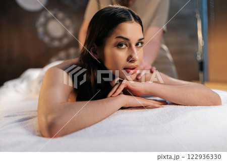 Portrait of beautiful young woman enjoying moment of wellness and pampering, getting massage in beauty parlor. Masseuse giving relaxing back massage to female client, looking at camera. Portrait of beautiful young woman enjoying moment of wellness and pampering, getting massage in beauty parlor. Masseuse giving relaxing back massage to female client, looking at camera. 122936330