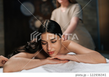 Closeup portrait of tranquil young woman lying on massage table with closed eyes during professional relaxing massage at spa salon. Professional massage therapist performing back treatment. 122936341