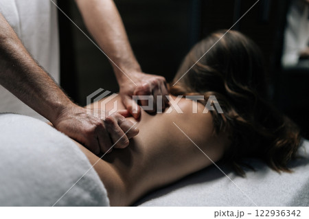 Close-up hands of masseur male performing deep tissue technique on female back, targeting muscle tension and promoting wellness during bodywork session in beauty parlor with dark interior. Close-up hands of masseur male performing deep tissue technique on female back, targeting muscle tension and promoting wellness during bodywork session in beauty parlor with dark interior. 122936342