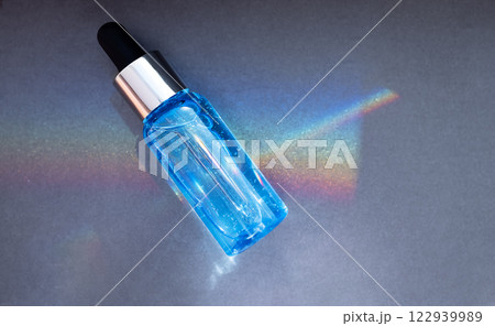 A close-up of a blue serum flask reflects a rainbow on a dark background. A mockup of a cosmetic product. A glass bottle with a pipette and moisturizing essence for the skin. 122939989