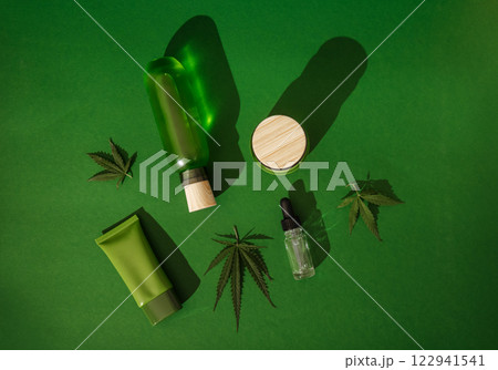 Clean mockups of cosmetic products and cannabis leaves. A glass bottle, a dropper, a cream package, a jar with a wooden lid on a green background. Clean mockups of cosmetic products and cannabis leaves. A glass bottle, a dropper, a cream package, a jar with a wooden lid on a green background. 122941541