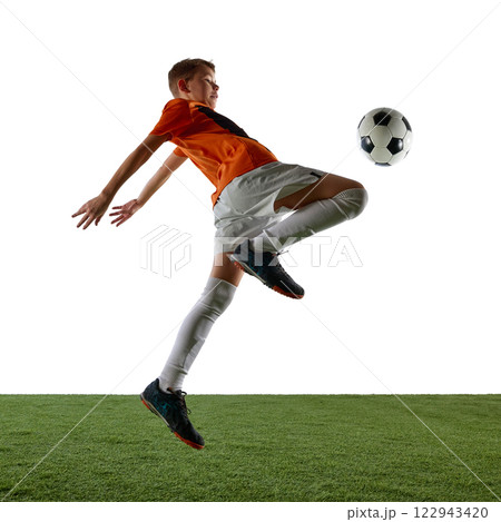 Dynamic image of focused boy in orange jersey and white shorts in motion, kicking ball with knee, training against white background 122943420