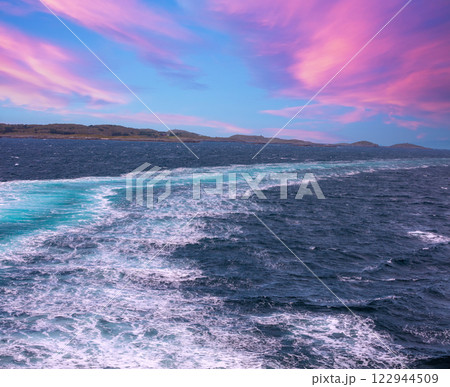 Island on the horizon. Rocks in the sea. Beautiful rocky sea landscape with dramatic cloudy sky. Water trail foaming behind a ferry. View from the ship. Nature of Norway 122944509