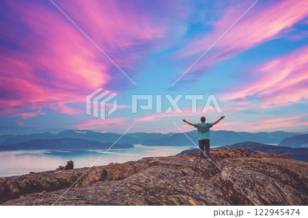 Panoramic view of the fjord. Twilight with a pink sky. A young man with hands in the air standing on a cliff of rock 122945474