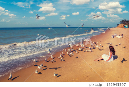 Seascape on a sunny day. A young woman sits on the beach, Seagulls are flying on the beach. The woman looks at the sea Seascape on a sunny day. A young woman sits on the beach, Seagulls are flying on the beach. The woman looks at the sea 122945597