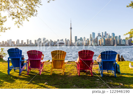 Colorful adirondack chairs on Toronto Island Park at sunset time. Toronto City downtown skyline in the background. Ontario, Canada. Colorful adirondack chairs on Toronto Island Park at sunset time. Toronto City downtown skyline in the background. Ontario, Canada. 122946190