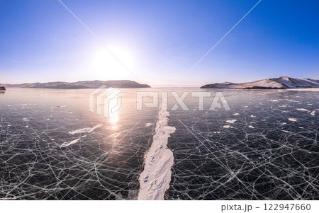 Aerial view on transparent frozen water, violet sunset in background. Beautiful winter landscape with clear blue smooth ice with cracks and mountains. Small Sea, Olkhon, Baikal lake, Siberia, Russia. 122947660