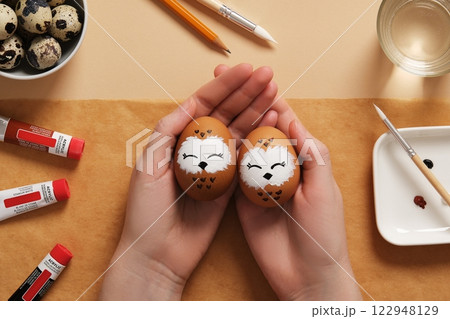 Hands holding Easter eggs with cute owls, hand-painted with acrylic paints, on a background of paints, brushes and palette. DIY concept 122948129