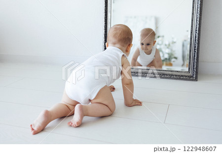 Sweet baby in white bodyswuit crawls across the floor to the mirror. Reflection of cute infant in mirror with beautiful frame. 122948607