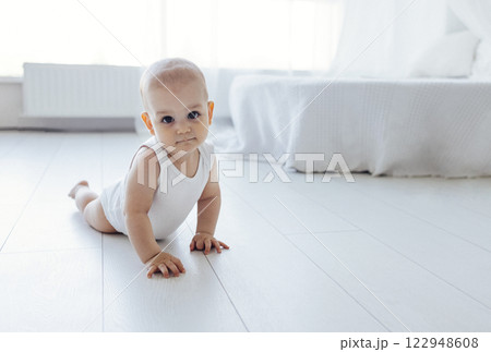 Sweet baby crawls on the floor of the bedroom. Cute little child in white bodysuit looks at the camera. 122948608