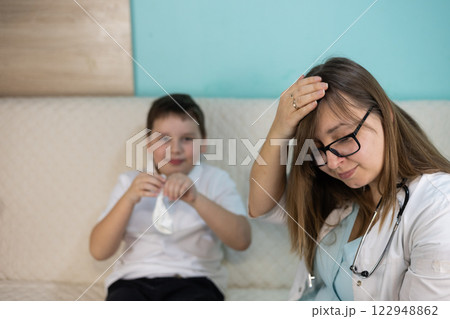 A tired pediatrician explains to a smiling child how to properly use a nasal blower at home to maintain the health of the eardrum. The doctor examines the child at home. A tired pediatrician explains to a smiling child how to properly use a nasal blower at home to maintain the health of the eardrum. The doctor examines the child at home. 122948862