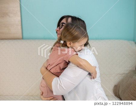 Female doctor wearing a white coat embracing a sick little girl during a home visit, offering comfort and care in a warm, familiar environment filled with empathy and support. Female doctor wearing a white coat embracing a sick little girl during a home visit, offering comfort and care in a warm, familiar environment filled with empathy and support. 122948894