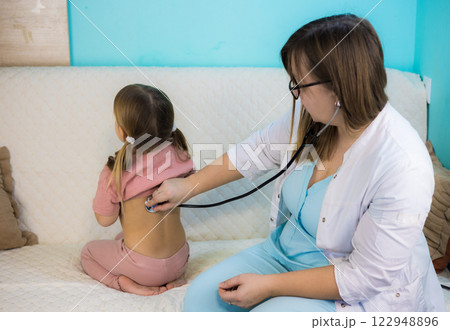 A doctor examines a sick child at home. A pediatrician uses a stethoscope to examine the lungs of a sick little girl sitting on a couch during a home visit to provide medical care. 122948896
