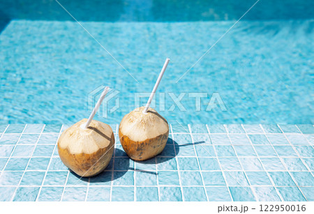 Close-up of two coconuts with straws on the side of the pool. Delicious natural cocktails on the background of clear blue water. Bright sunlight and tropical drinks. 122950016