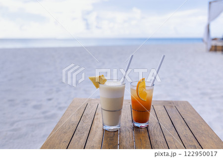 Two cocktails with slices of fruit are on a wooden table on the beach. Delicious chilled drinks on the background of tropical trees and the ocean. Two cocktails with slices of fruit are on a wooden table on the beach. Delicious chilled drinks on the background of tropical trees and the ocean. 122950017