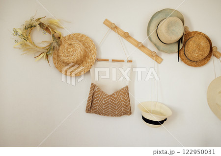 A white wall with wooden hangers with various straw summer hats hanging. Cotton linen wall hanging storage bag and wreath. A white wall with wooden hangers with various straw summer hats hanging. Cotton linen wall hanging storage bag and wreath. 122950031