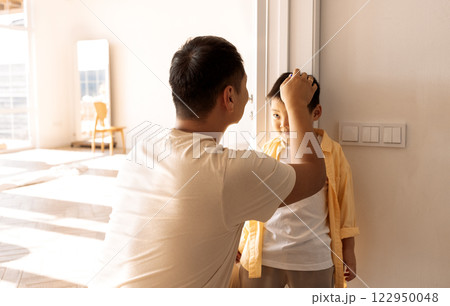 A young Asian man marks the height of his little son on the doorway in the living room. Dad and his cute boy are having fun at home together. 122950048