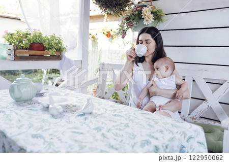 Young beautiful mother drinks tea and holds cute baby in white bodysuit in her arms. Young beautiful mother drinks tea and holds cute baby in white bodysuit in her arms. 122950062