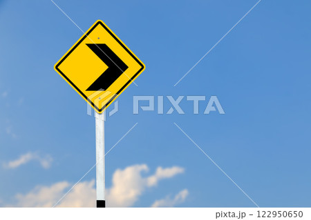 Black right direction symbol is displayed on a bright yellow traffic sign on blurred sky background. Black right direction symbol is displayed on a bright yellow traffic sign on blurred sky background. 122950650