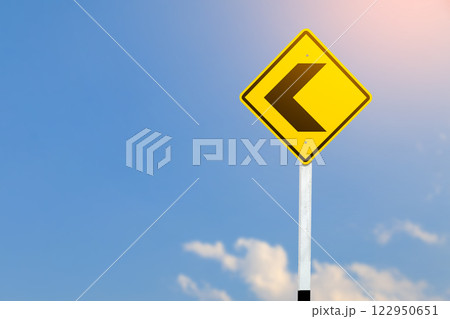 Black left direction arrow is displayed on a bright yellow traffic sign on blurred sky background. Black left direction arrow is displayed on a bright yellow traffic sign on blurred sky background. 122950651