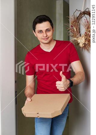 positive delivery man in red t-shirt giving food order, holding pizza box standing at doorway 122951145