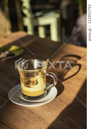 Glass cup with hot golden turmeric latte on wooden table in sunlight. Warm autumn mood, cozy coffee shop atmosphere Glass cup with hot golden turmeric latte on wooden table in sunlight. Warm autumn mood, cozy coffee shop atmosphere 122951752
