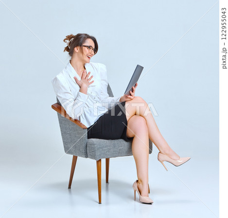 Young woman sitting on chair, wearing glasses, holding tablet, smiling warmly, engaged in conversation against light blue studio background. Young woman sitting on chair, wearing glasses, holding tablet, smiling warmly, engaged in conversation against light blue studio background. 122955043