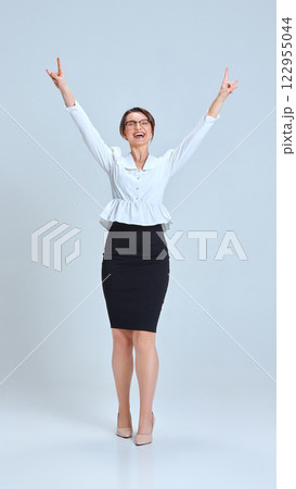 Young woman in white blouse and black skirt raising hands, celebrating victory, smiling brightly against light blue studio background. Young woman in white blouse and black skirt raising hands, celebrating victory, smiling brightly against light blue studio background. 122955044