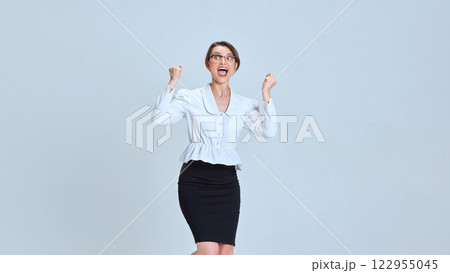Young woman raising fists in air, wearing glasses and formal outfit, expressing excitement against light blue studio background. Young woman raising fists in air, wearing glasses and formal outfit, expressing excitement against light blue studio background. 122955045