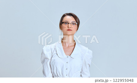 Serious young woman, in white blouse and glasses, maintaining professional posture against light blue studio background. Serious young woman, in white blouse and glasses, maintaining professional posture against light blue studio background. 122955048