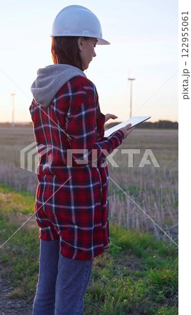 Woman engineer wearing a white protective helmet is taking notes with a clipboard in a field with wind turbines, as the sun sets. Clean energy and engineering concept Woman engineer wearing a white protective helmet is taking notes with a clipboard in a field with wind turbines, as the sun sets. Clean energy and engineering concept 122955061