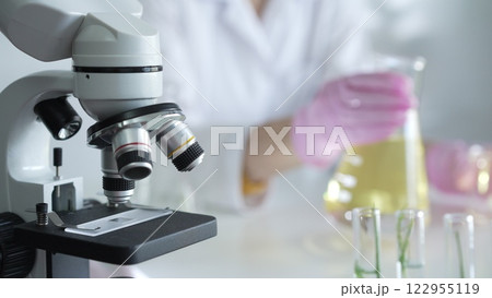 Microscope is near a scientist, wearing a lab coat and pink protective gloves, and holding beaker with a yellow oily liquid in laboratory setting, close up. Medicine and science Microscope is near a scientist, wearing a lab coat and pink protective gloves, and holding beaker with a yellow oily liquid in laboratory setting, close up. Medicine and science 122955119