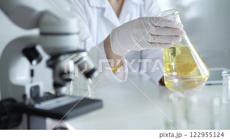 Microscope is near a scientist, wearing a lab coat and white protective gloves, and holding beaker with a yellow oily liquid in laboratory setting, close up. Medicine and science concept Microscope is near a scientist, wearing a lab coat and white protective gloves, and holding beaker with a yellow oily liquid in laboratory setting, close up. Medicine and science concept 122955124