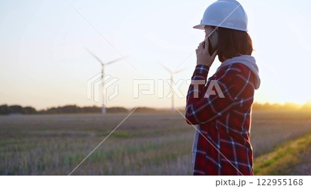 Woman engineer talking on the phone in a field with wind turbines, as the sun sets. Concept of clean energy and engineering 122955168
