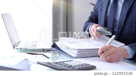 Businessman using a calculator and magnifying glass while analyzing financial documents at desk in fair modern office. Audit and taxes in business 122955226