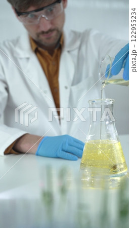 Male scientist in lab coat, blue gloves, and protective glasses, is pouring a yellow oily liquid from one beaker to another in a laboratory, vertical close-up view. Science and medicine concepts 122955234