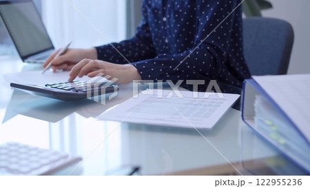A female accountant with blue dotted blousy is using a laptop computer and calculator to calculate taxes at a glass desk in the office. Left side view A female accountant with blue dotted blousy is using a laptop computer and calculator to calculate taxes at a glass desk in the office. Left side view 122955236