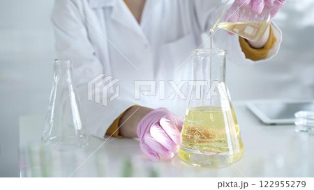 A scientist, wearing a lab coat and pink protective gloves, is pouring a yellow oily liquid from one beaker to another in laboratory, close up. Medicine and science 122955279