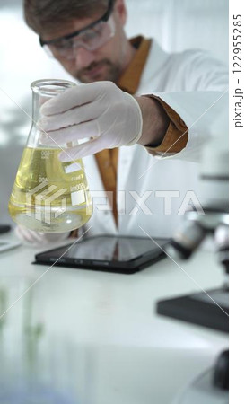 Man scientist wearing white protective gloves, and glasses is holding a yellow chemical solution inside an Erlenmeyer flask in a laboratory, vertical portrait 122955285