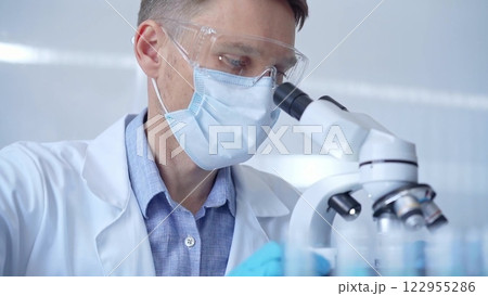 Man scientist with protective glasses, mask and gloves is using microscope in laboratory, portrait of researcher. Microbiology science concept Man scientist with protective glasses, mask and gloves is using microscope in laboratory, portrait of researcher. Microbiology science concept 122955286