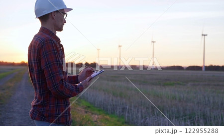 Man engineer wearing a white protective helmet is taking notes with a clipboard in a field with wind turbines, as the sun sets. Clean energy and engineering concept 122955288